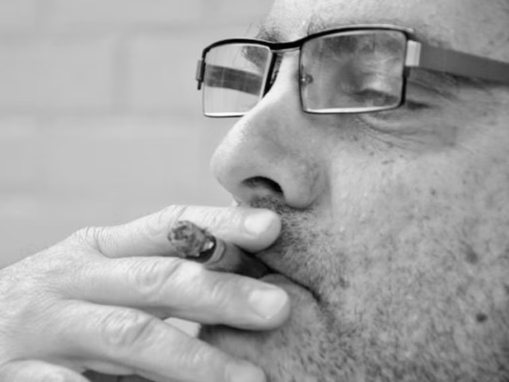Homem de óculos e barba fumando um cigarro.